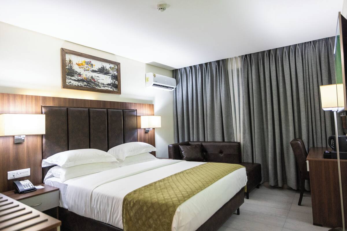 A hotel room with a large bed, a desk, a chair, a nightstand, a lamp, an air conditioner, and a painting on the wall.
