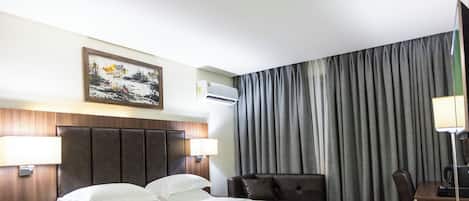 Ein Hotelzimmer mit einem großen Bett, einem Schreibtisch, einem Sessel, einem Nachttisch, einer Lampe, einer Klimaanlage und einem Gemälde an der Wand.