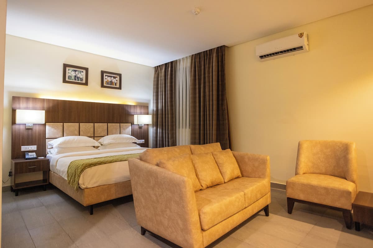 A hotel room with a bed, a sofa, a chair, an air conditioning unit, and a nightstand.