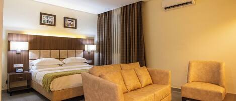 A hotel room with a bed, a sofa, a chair, an air conditioning unit, and a nightstand.