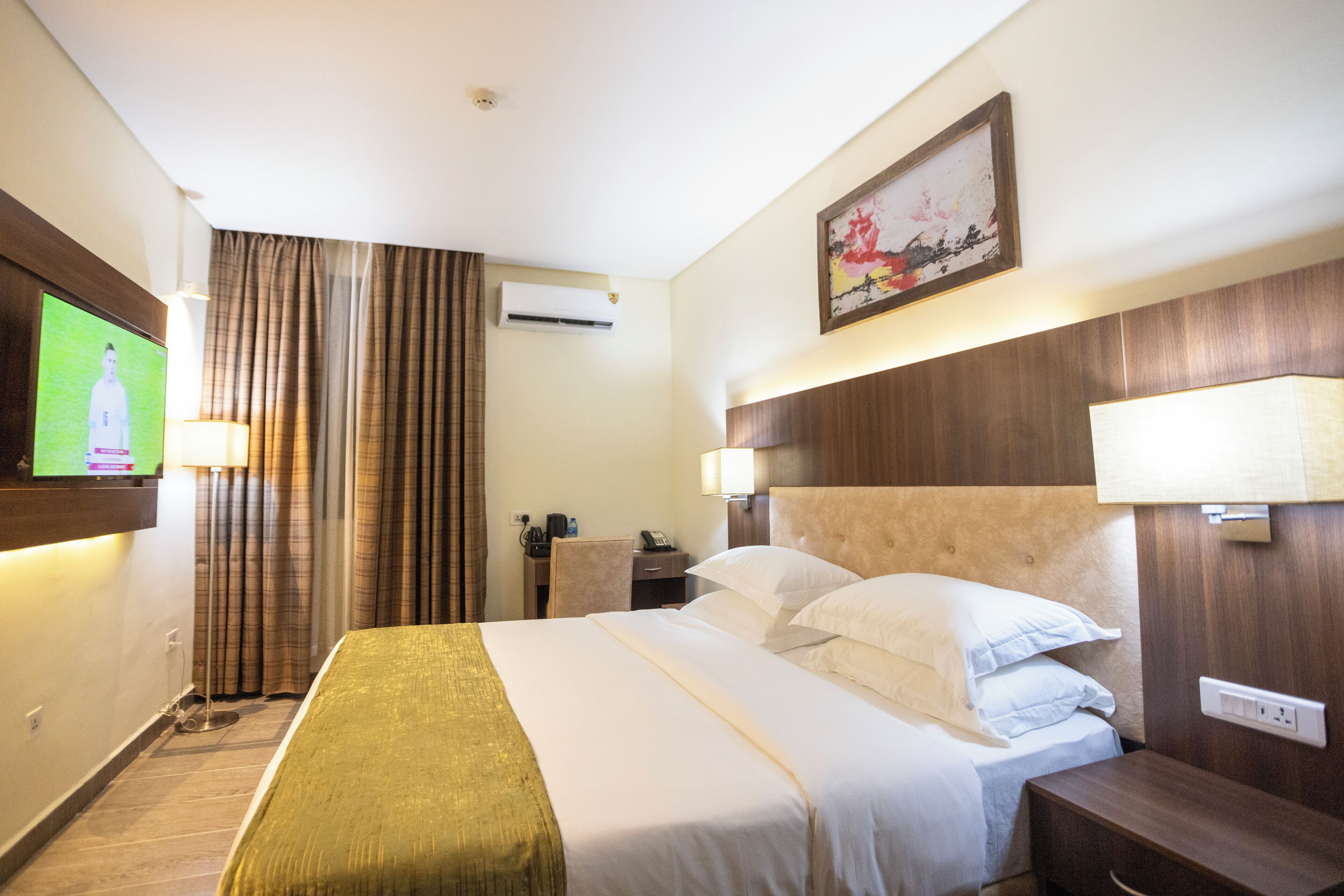 A hotel room with a large bed, a TV mounted on the wall, a desk with a lamp, and a painting above the bed.