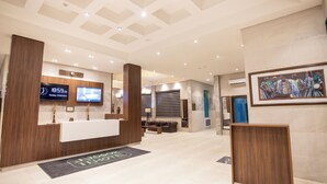 Zona de recepción de un hotel, con mostrador de recepción, relojes digitales y una obra de arte enmarcada en la pared.