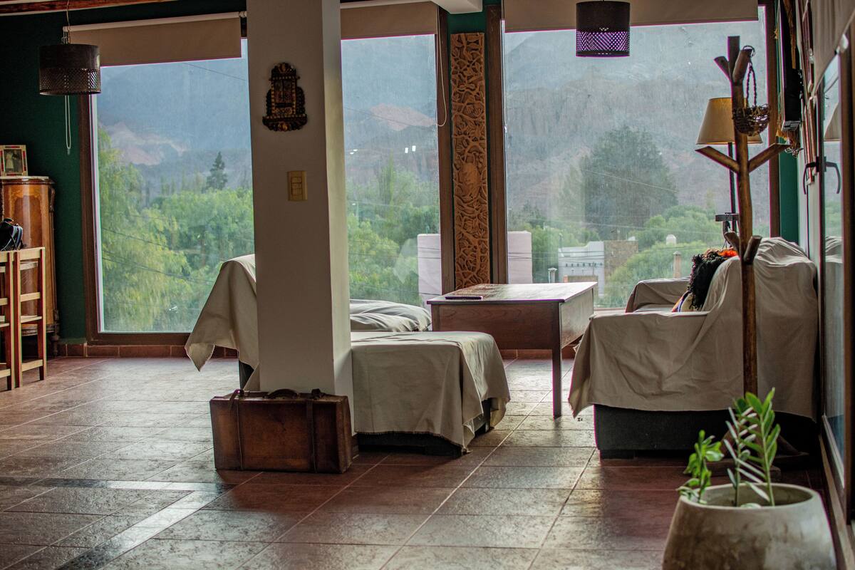 A room with a large window, a bed, a table, and a chair. There is a suitcase on the floor and a plant on the table.