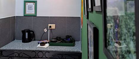 A small kitchenette with a refrigerator, a kettle, and a microwave. There is a window with a view of trees and a building.