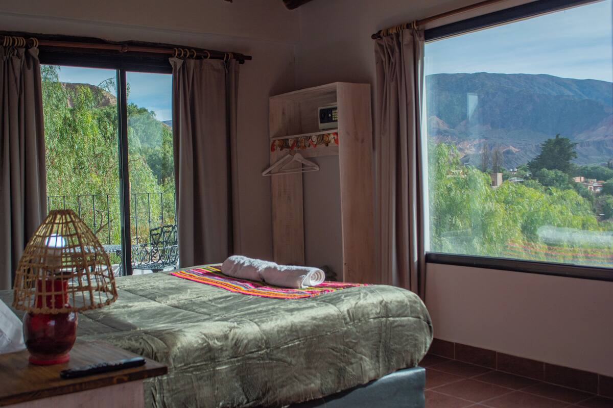 A bedroom with a large window, a bed, a hanging clothes rack, and a table with a lamp.