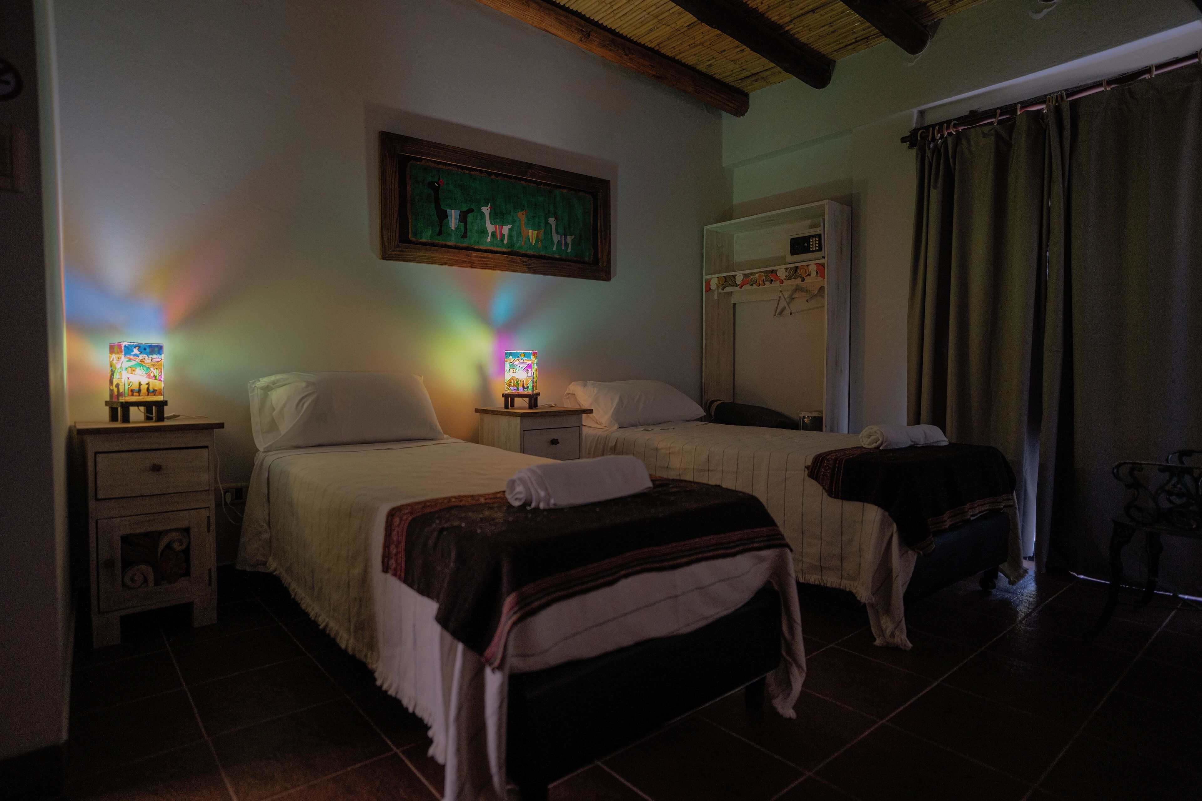 Habitación de hotel con dos camas, una mesita de noche con lámpara, un cuadro en la pared y una ventana con cortinas.