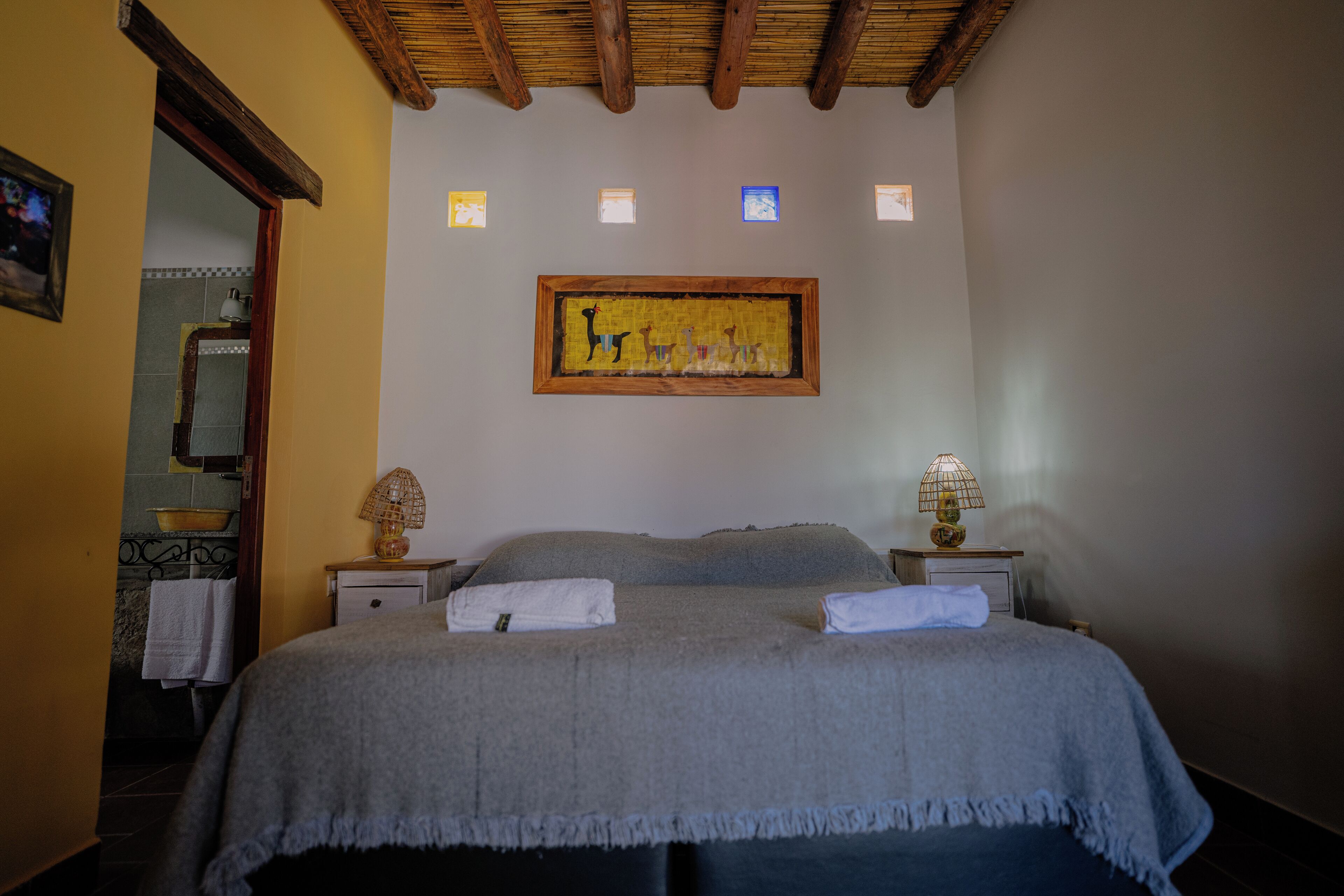 A bedroom with a bed, two bedside tables, a lamp, a framed picture on the wall, and a bathroom visible through an open door.