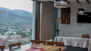 Una habitación con una mesa de madera, sillas, una cama con cubrecama blanco, un televisor montado en la pared y vistas a montañas y edificios a través de la ventana.