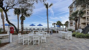 Un espace extérieur aménagé en terrasse, avec des chaises et des tables blanches, des palmiers et des parasols bleus.