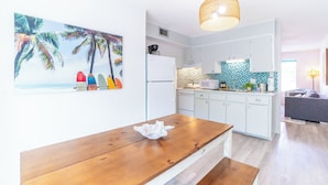 A modern kitchen with white cabinets, a wooden dining table, and a beach-themed wall art. -  Sports Weekend Stay Near Fields, Beach & Pool onsite. Coffee/Wine in backyard (Gulf Shores)