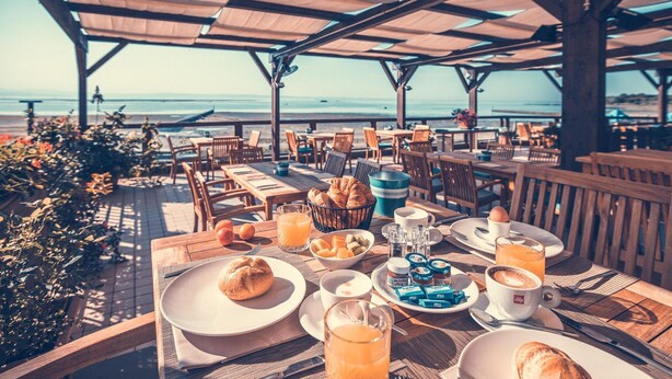 A breakfast table set on a wooden deck with a view of the ocean, featuring croissants, fruit, and coffee. - Lodge Tenuta Primero in Grado (Province of Gorizia)