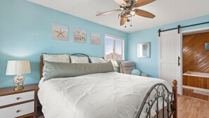 Une chambre à coucher comprenant un lit, une table de chevet, une lampe, un ventilateur de plafond et des œuvres d’art accrochées aux murs.