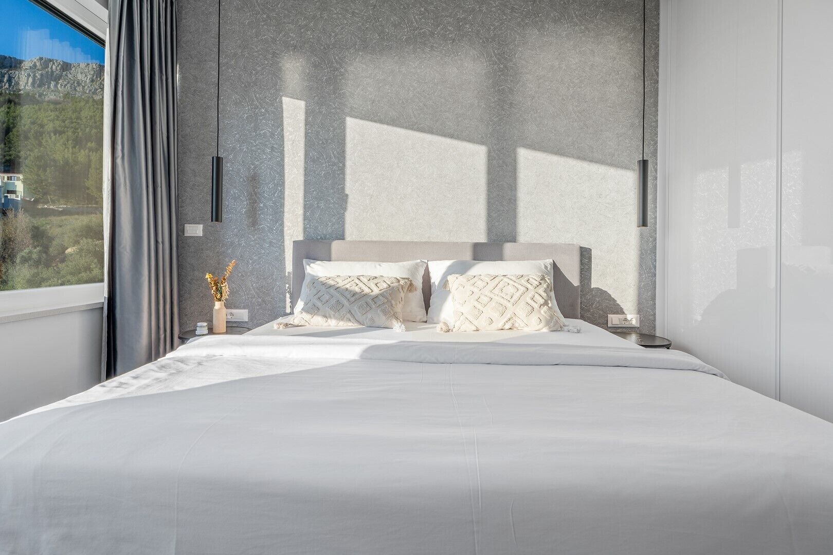 A modern bedroom with a large bed, a nightstand, and a view of a mountainous landscape through a window.