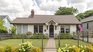 Une maison de plain-pied avec un porche à l’avant, une cheminée et un petit jardin agrémenté de fleurs blanches.