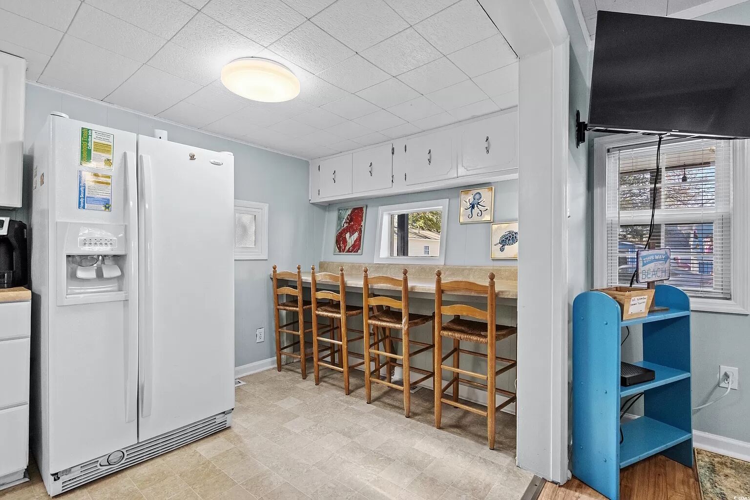 A kitchen with a white refrigerator, wooden chairs, a blue shelf, and a window with a beach sign.