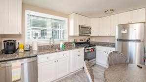A modern kitchen with white cabinets, a stainless steel refrigerator, a microwave, a stove, and a dining area with a chair and table.