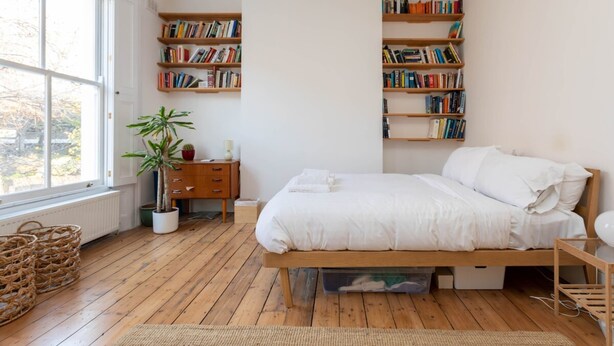 A bedroom with a wooden bed, a bedside table, a nightstand, a potted plant, and built-in bookshelves. - Spacious 2BD Flat By Regents Park & Camden Town! (London)