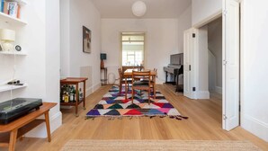 A spacious living area with a wooden floor, a colorful rug, a dining table with chairs, a bookshelf, and a piano. - Spacious 2BD Flat By Regents Park & Camden Town! (London)