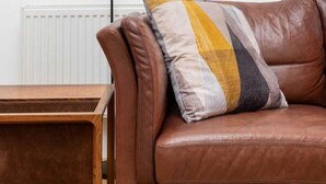 A brown leather sofa with a multicolored pillow, a wooden side table, and a floor lamp against a white wall. - Spacious 2BD Flat By Regents Park & Camden Town! (London)