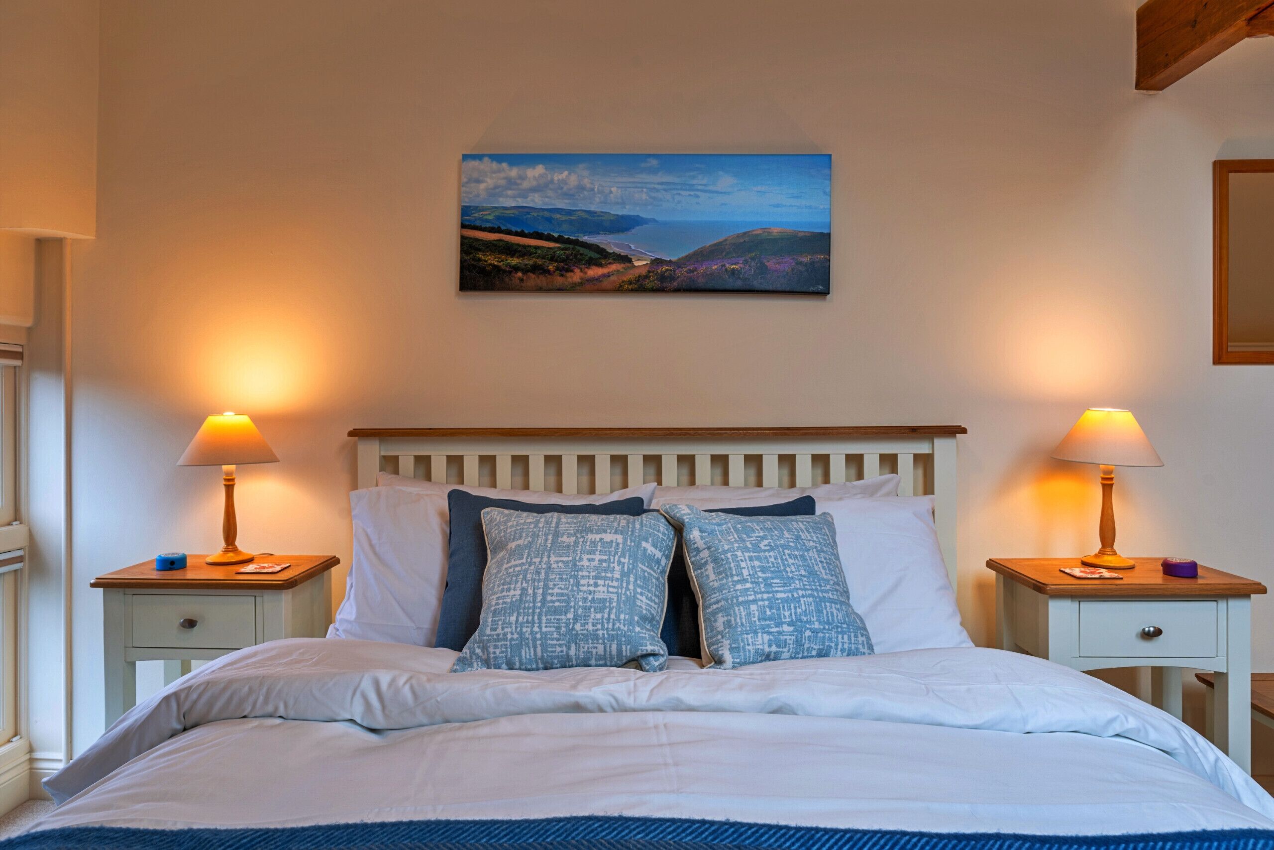 A bedroom with a bed, two bedside tables with lamps, a painting on the wall, and a wooden headboard.