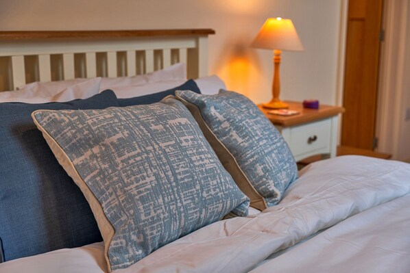 A neatly made bed with patterned pillows, a wooden headboard, a bedside lamp, and a small wooden nightstand with a lamp and a purple object. - Lower School Cottages (Minehead)