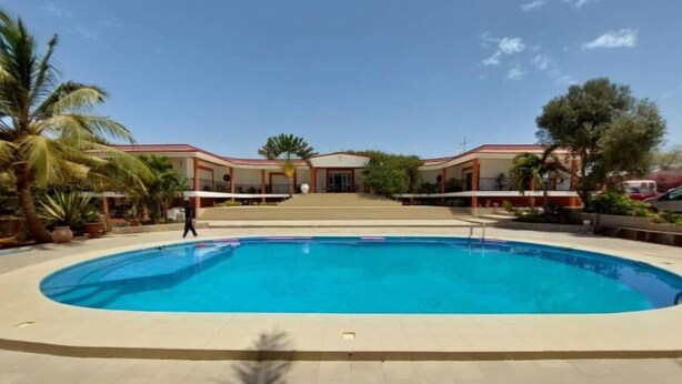 Una piscina con bordo piastrellato, palme e un edificio con tetto rosso sullo sfondo.