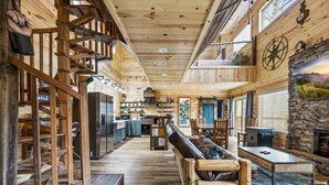 Un interior espacioso con una escalera de madera, un sofá de cuero, una chimenea de piedra y una zona de cocina.