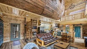 Interior espacioso de una cabaña de madera con una sala de estar acogedora que incluye un sofá de cuero, una mesa de comedor de madera y una cocina al fondo.