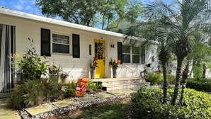 A white house with a yellow door, surrounded by a garden with palm trees and various plants. - South Tampa! Stay Steps from Bayshore Near MacDill AFB (Tampa)
