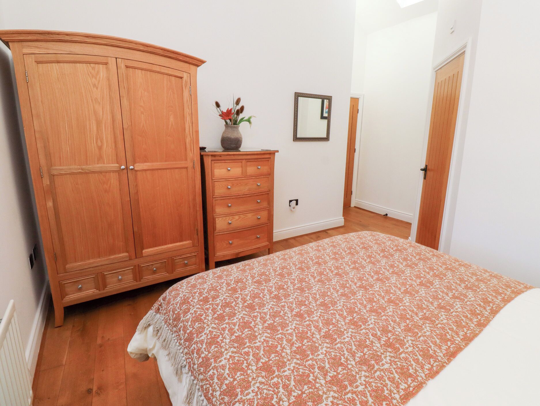 A bedroom with a large wooden wardrobe, a dresser, a bed with a patterned blanket, a vase with flowers, and a framed mirror on the wall.