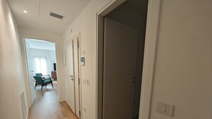 En smal hall med trägolv, vita väggar och en dörr som leder in till ett rum. Längst bak i hallen står en stol och ett bord, och på väggen sitter en ljusströmbrytare.