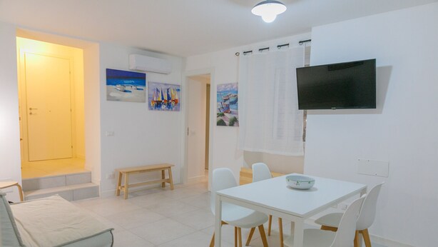 A bright, modern living room with a white sofa, a dining table with chairs, a flat-screen TV mounted on the wall, and artwork on the walls.