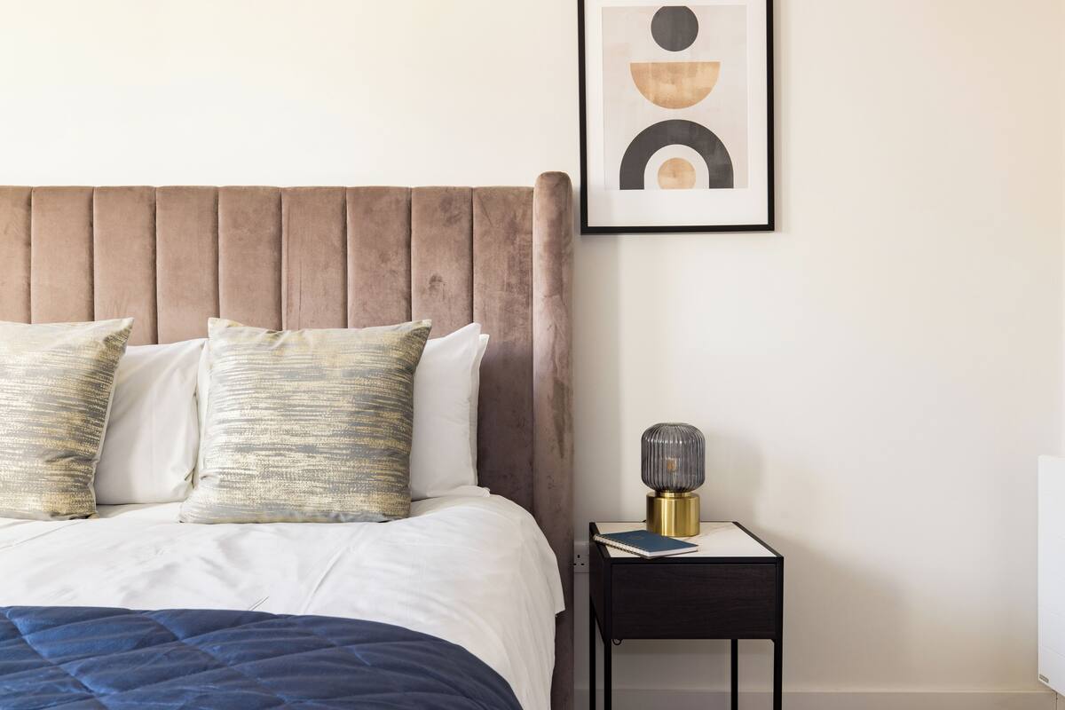A neatly made bed with a blue quilt, a wooden headboard, and two decorative pillows. A bedside table with a lamp and a book is visible.
