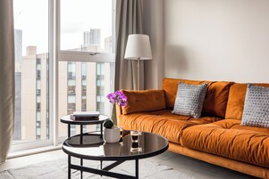 A modern living room with a sofa, a coffee table, a lamp, and a view of the cityscape through large windows.