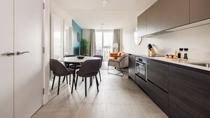 Une cuisine moderne avec des meubles de couleur sombre, un coin repas avec des chaises et une table, et une vue sur le salon à travers une porte ouverte.