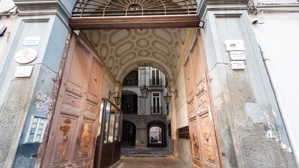 Una entrada apuntada amb portes rovellades i un camí de pedra que condueix a un edifici amb un balcó.