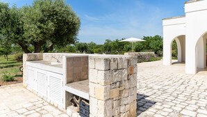Une structure en pierre située à l’extérieur, dotée d’un barbecue intégré, entourée d’une zone pavée et d’un bâtiment blanc orné d’arcades.