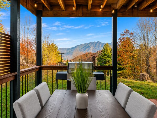 An outdoor patio with a table and chairs, a grill, and a view of mountains and autumn trees.