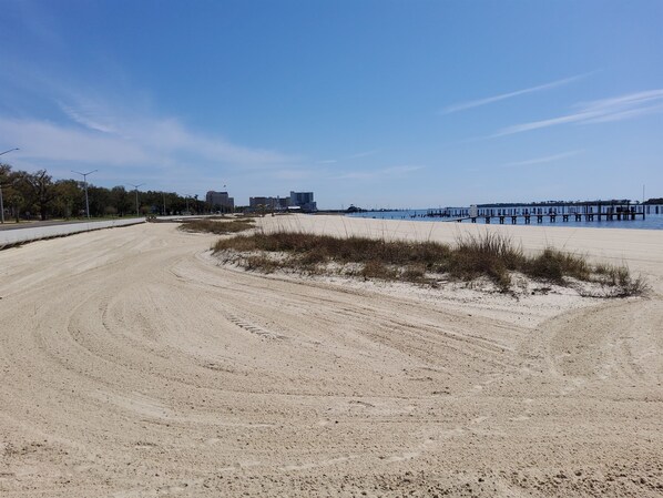 Una playa arenosa con un muelle que se adentra en el agua, edificios al fondo y un cielo despejado.