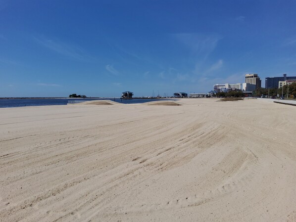 A wide sandy beach with a clear blue sky and calm sea. There are buildings in the background.