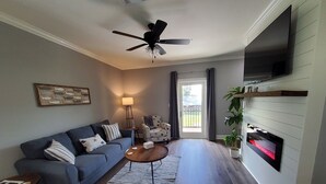A living room with a blue sofa, a coffee table, a TV mounted on the wall, and a ceiling fan. - Four Palms Unit D, 500ft to Beach, Walk 2 Casinos! (Biloxi)