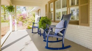 A covered porch with outdoor seating, potted plants, and a view of a garden with pink flowers. - Pet friendly, huge fenced in yard, Fireplace, Firepit, near SGMC & VSU & Eats (Valdosta)