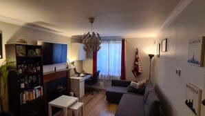 Ein Wohnzimmer mit einer Couch, einem Fernseher, einem Schreibtisch und einem Bücherregal.