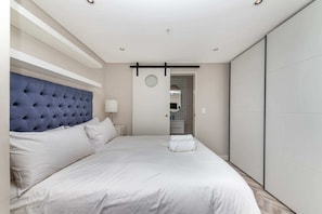Un dormitorio moderno con un cabecero capitonado, una cama grande con ropa blanca, una mesita de noche con lámpara y un armario con puerta corredera.