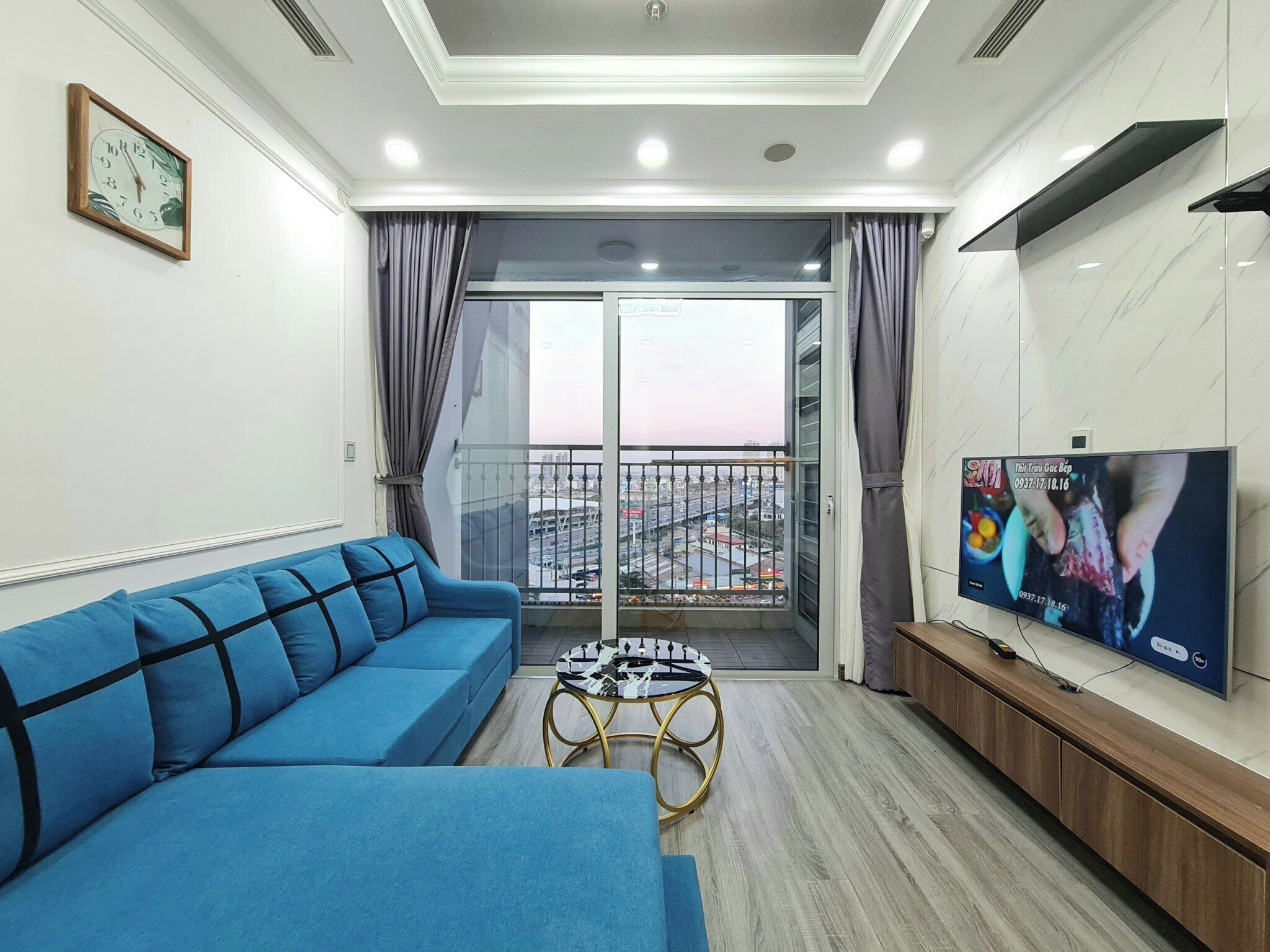 A modern living room with a blue sofa, a small round coffee table, a TV on the wall, and a balcony with a city view.