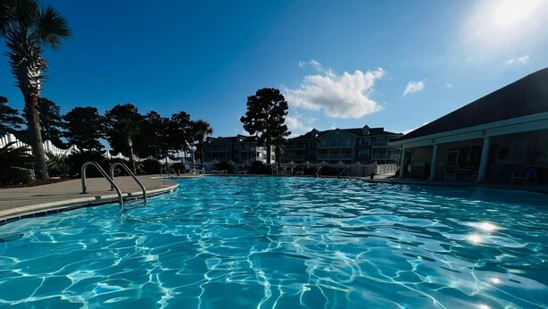 A clear blue swimming pool with a metal ladder, surrounded by palm trees and a building with multiple windows in the background. - Beaches and Birdies II -  First Floor Golf Resort Studio (Calabash)