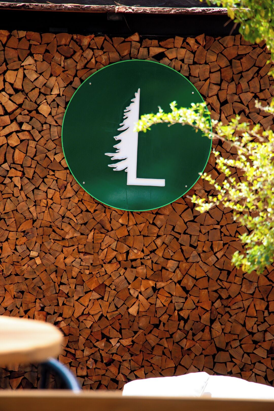 A circular sign with a white 'L' on a green background, mounted on a wall covered with stacked wood logs.