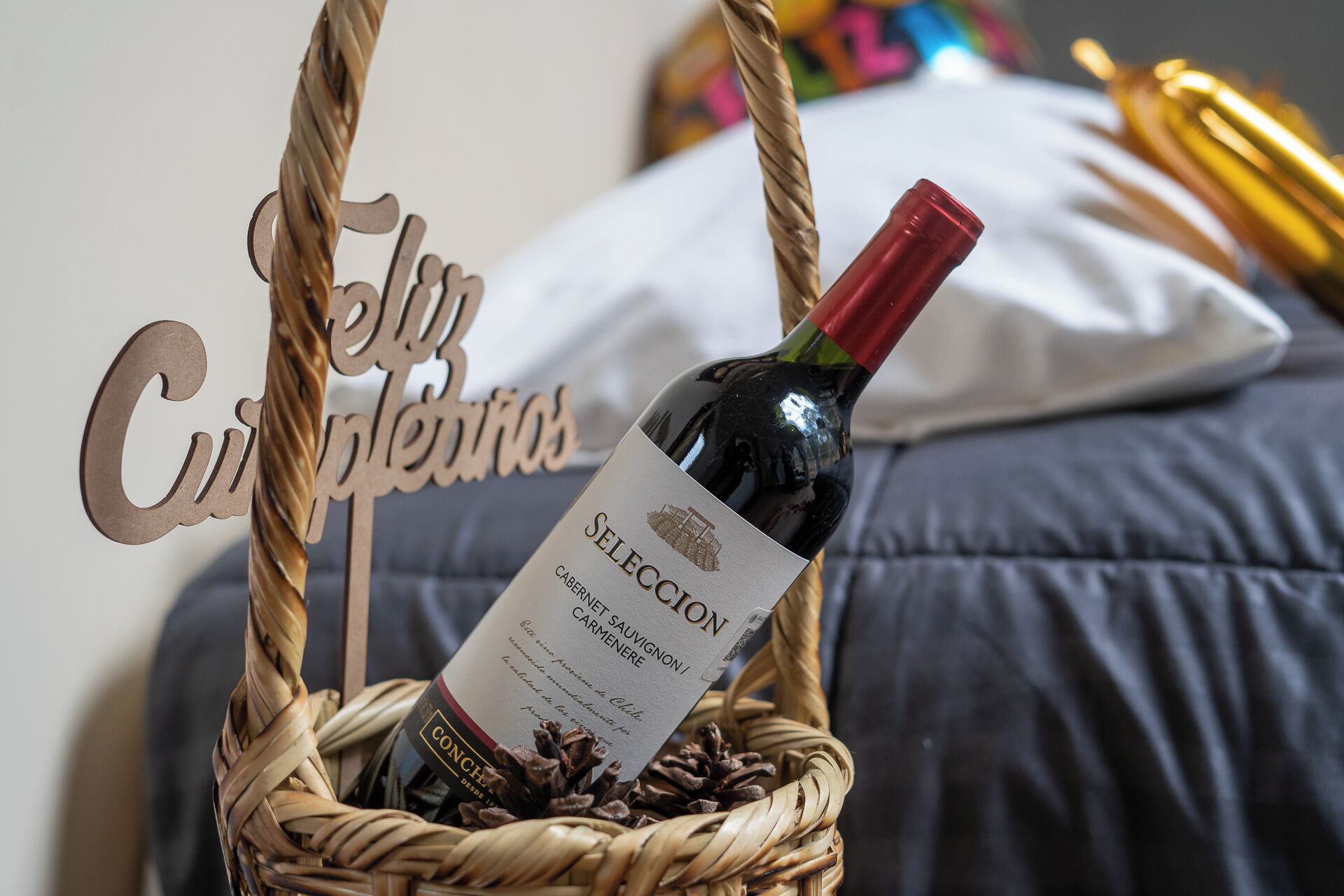 A bottle of wine in a woven basket with pine cones, placed on a bed with a pillow and a decorative sign.