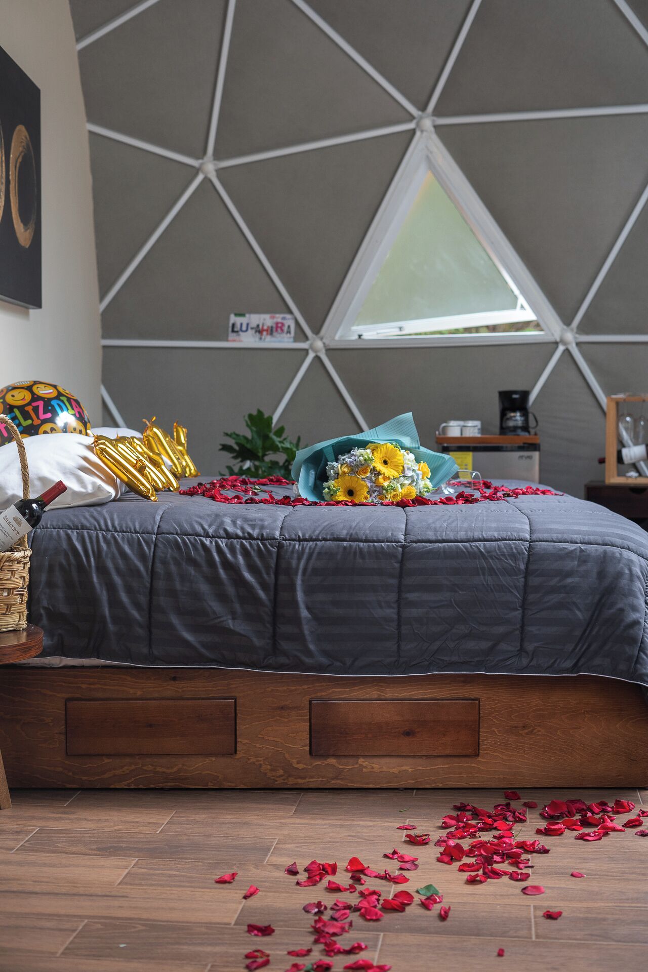 Ein Schlafzimmer mit einer deckenhohen Wandgestaltung im geometrischen Muster, einem Bett mit grauer Decke, einem Korbwannenkorb mit einer Weinflasche, einem Blumenstrauß und verstreuten Rosenblättern auf dem Boden.