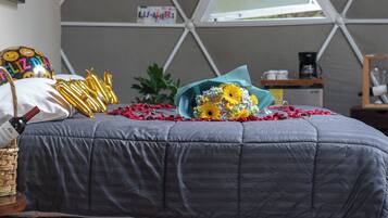 Ein Schlafzimmer mit einer deckenhohen Wandgestaltung im geometrischen Muster, einem Bett mit grauer Decke, einem Korbwannenkorb mit einer Weinflasche, einem Blumenstrauß und verstreuten Rosenblättern auf dem Boden.